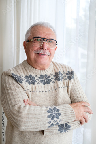 Happy and smile Senior man in glasses 70-75 years old, standing by window with armcross on breast and looking to camera