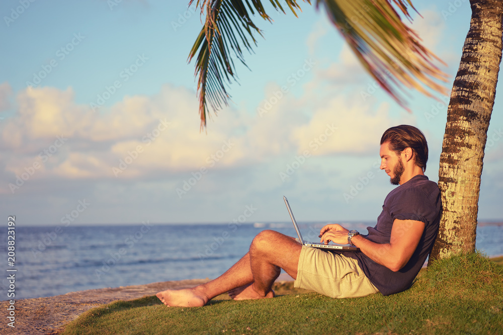 Work and vacation. Young man working on laptop computer on the tropical beach under the palm tree.