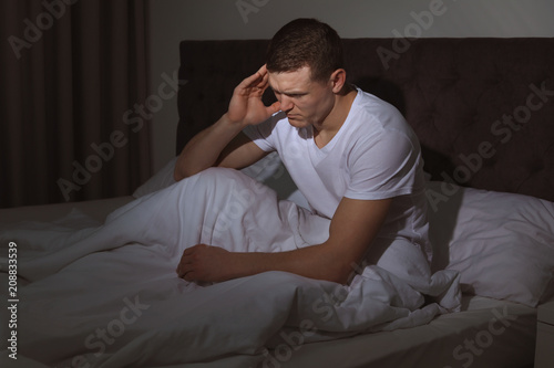 Handsome young man suffering from headache while sitting in bed at night