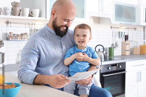 Dad reading newspaper while holding little son in kitchen