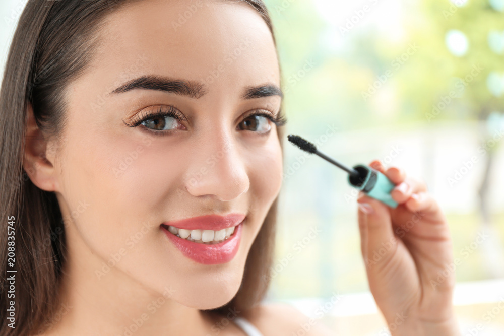Attractive young woman applying mascara on her eyelashes against blurred background Attractive young woman applying mascara on her eyelashes against blurred background