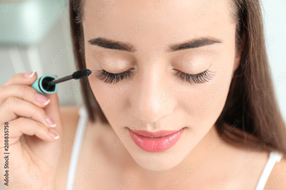 Attractive young woman applying mascara on her eyelashes against blurred background Attractive young woman applying mascara on her eyelashes against blurred background