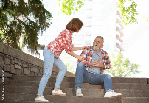 Woman helping mature man suffering from heart attack on stairs, outdoors