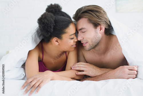 portrait of multiethnic young couple in love lying under blanket together in bed in morning