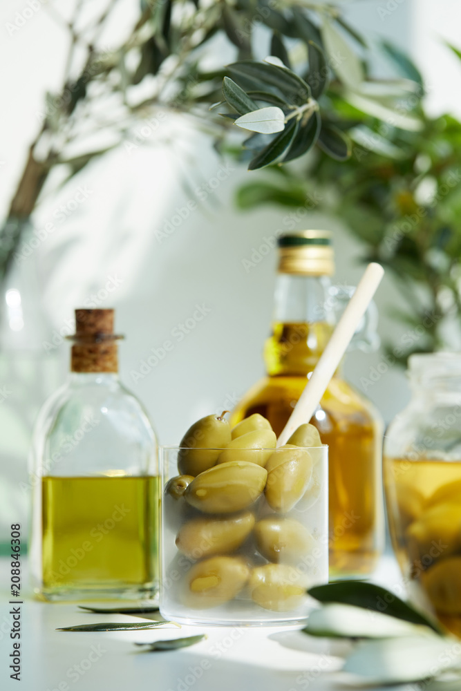 glass with spoon and green olives, jar, various bottles of aromatic olive oil with and branches on white table glass with spoon and green olives, jar, various bottles of aromatic olive oil with and branches on white table
