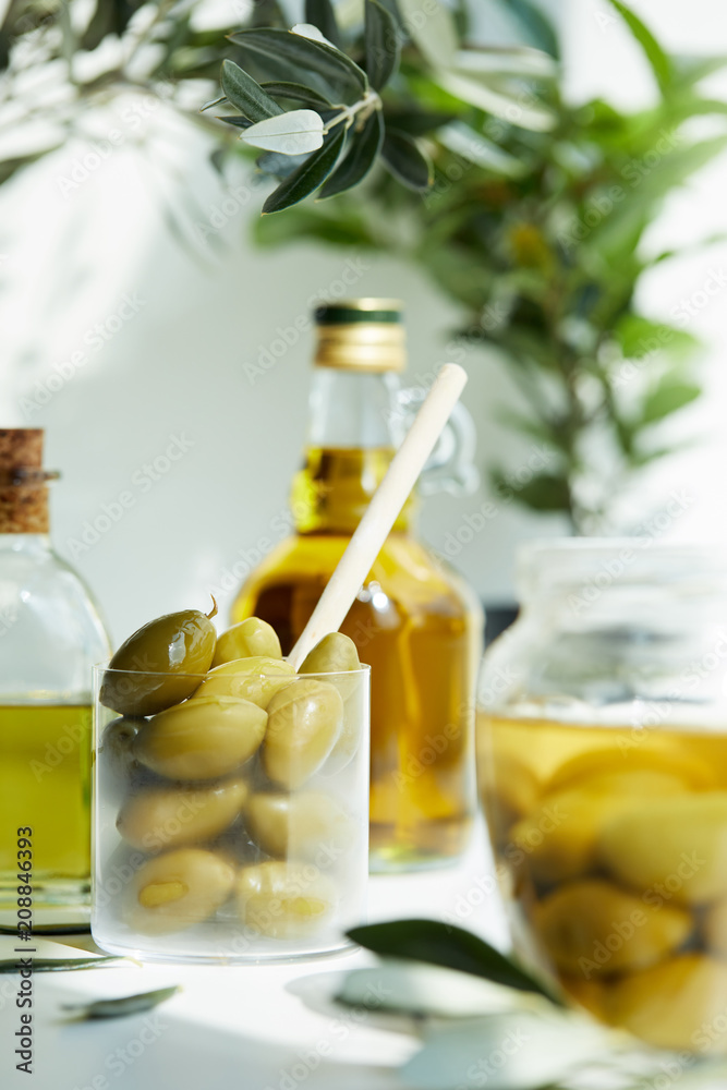 glass with spoon and green olives, jar, various bottles of aromatic olive oil with and branches on white table glass with spoon and green olives, jar, various bottles of aromatic olive oil with and branches on white table