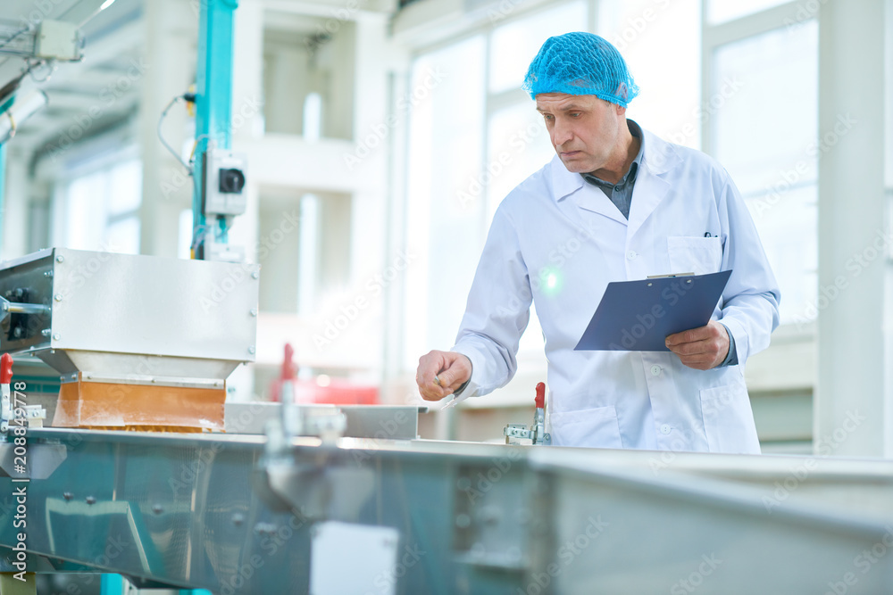 Waist up portrait of senior factory worker doing production quality inspection holding clipboard and making notes standing by conveyor belt in modern production workshop, copy space Waist up portrait of senior factory worker doing production quality inspection holding clipboard and making notes standing by conveyor belt in modern production workshop, copy space