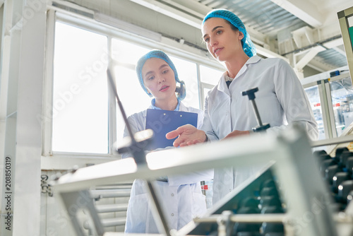 Low angle portrait of  two young female workers wearing lab coats standing by  power units and writing on clipboard  in clean production workshop  copy space