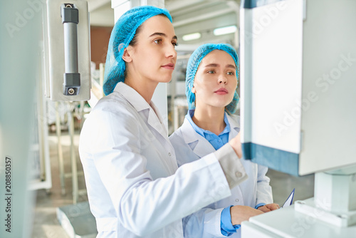 Waist up portrait of two young female workers wearing lab coats standing by power units and pressing buttons on control panel in clean production workshop, copy space
