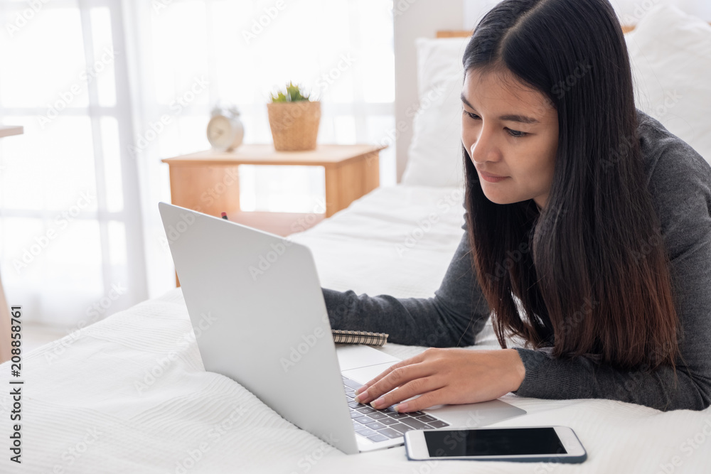 Asian woman laying on bed doing paperwork on laptop in bedroom.work at home.e-larning concept.study online. Asian woman laying on bed doing paperwork on laptop in bedroom.work at home.e-larning concept.study online.