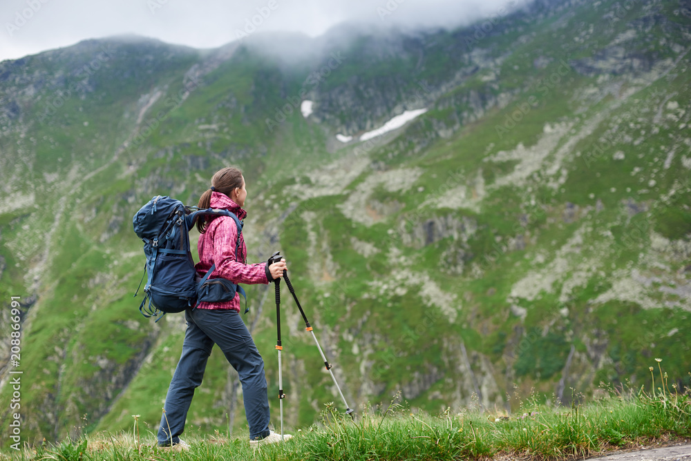 Hiker girl in modern outfit with blue backpack and trekking sticks walking in green rocky mountains with foggy clouds. Young hiker woman in pink jacket and gray pants enjoying view of high a mountain.