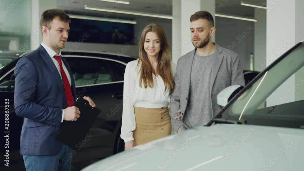 Friendly car salesman is talking to confident young man telling him about new vehicle model while pretty woman is standing near her husband holding his arm and smiling. Friendly car salesman is talking to confident young man telling him about new vehicle model while pretty woman is standing near her husband holding his arm and smiling.