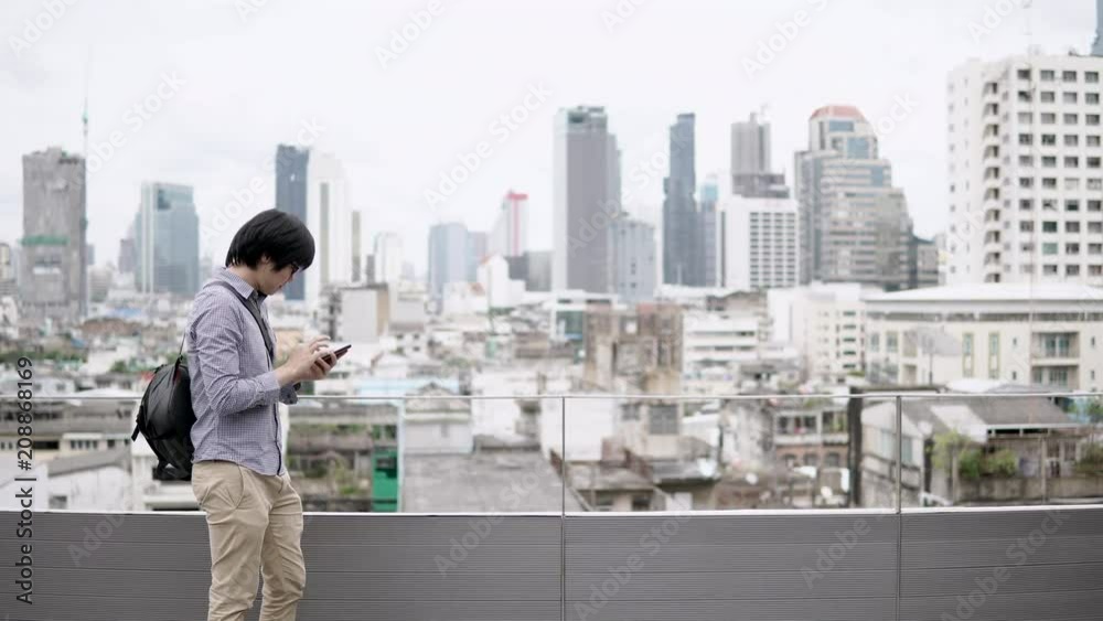 Young Asian businessman using smartphone on office building rooftop terrace with city view in the background. Using social media application or playing games on smart device concepts. 4K video Young Asian businessman using smartphone on office building rooftop terrace with city view in the background. Using social media application or playing games on smart device concepts. 4K video