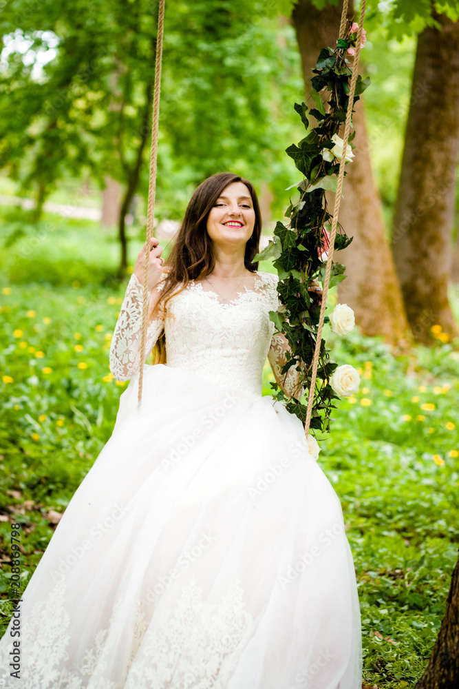 Beautiful bride is sitting on a swing decorated with flowers in the park