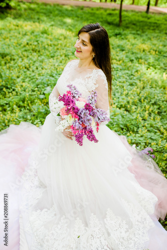 Amazing girl in a white dress sitting on the grass in a park with a bouquet in her hands