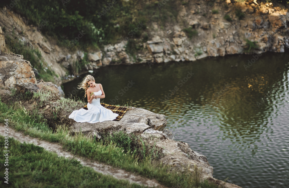 A beautiful bride with curly hair sits on a plaid, against the background of rocks and a river, and examines the flowers. Beautiful bride hair blows the wind.