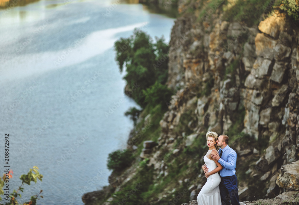 A stylish bridegroom embraces a beautiful bride from behind, against the background of huge rocks and the sea. Lovely newlyweds stand on a cliff. Wedding portrait of gentle newlyweds.