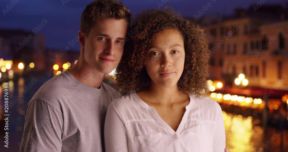 Millennial couple pose for a portrait by the Grand Canal in evening, Multiethnic male and female tourists in Venice smile at camera, 4k