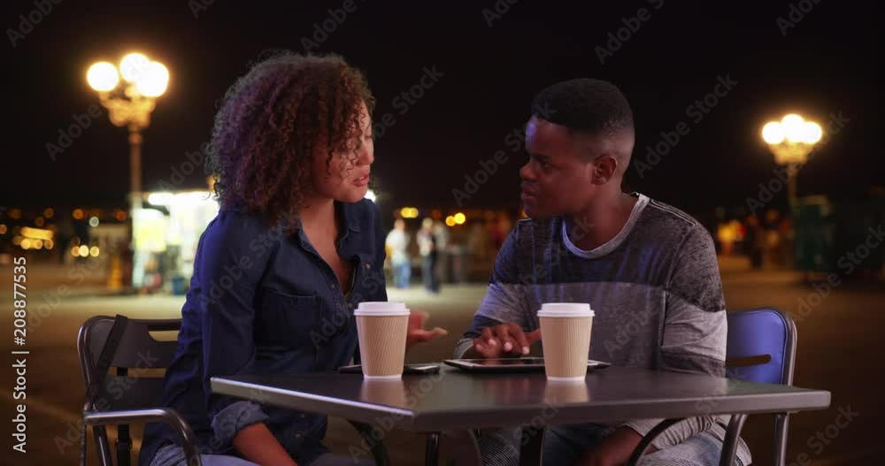 Black male and female sit outside at night using touchscreen devices, African American man and woman meeting for coffee, chat and use technology, 4k Black male and female sit outside at night using touchscreen devices, African American man and woman meeting for coffee, chat and use technology, 4k
