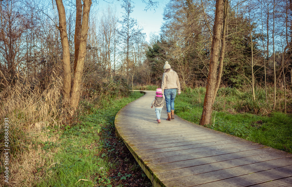 Back view of mother and daughter walking together holding hands over a wooden pathway into the forest