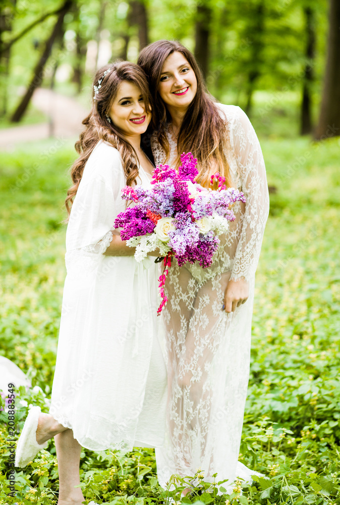Two cute brides are standing in the park with a bouquet in their hands Two cute brides are standing in the park with a bouquet in their hands