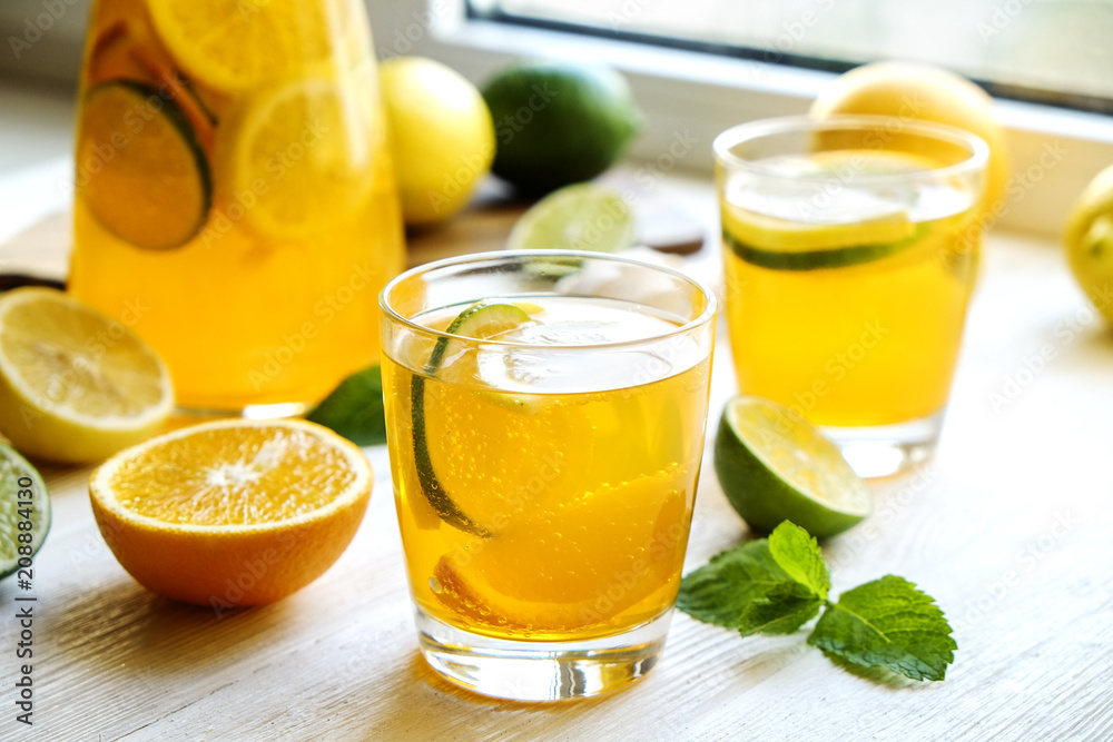 Iced lemonade pitcher & two glasses, wooden juicer, cold citrus infused water, lemon & lime, mint leaves, cutting board on white wooden windowsill. Apartment window background, close up, copy space.