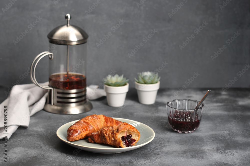 Two crispy croissants on a plate. One croissant is broken and smeared with jam. Next a cup of jam and a teapot with tea. In the frame, cloth napkin. Gray background. Close-up. Two crispy croissants on a plate. One croissant is broken and smeared with jam. Next a cup of jam and a teapot with tea. In the frame, cloth napkin. Gray background. Close-up.