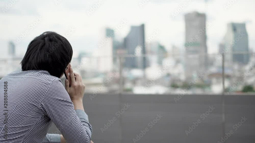 Young Asian businessman using smartphone for business talk sitting on office building rooftop terrace with city view in the background. Business communication and corporation concepts. 4K video Young Asian businessman using smartphone for business talk sitting on office building rooftop terrace with city view in the background. Business communication and corporation concepts. 4K video