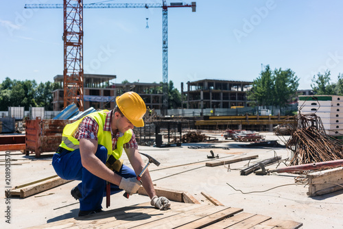 Fotografija Side view of a young worker wearing safety vest and yellow hard hat, while hamme