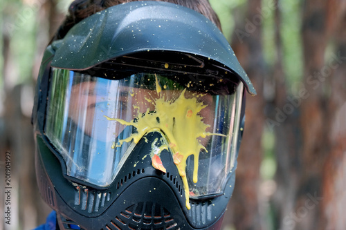 Yellow blot paint on a protective glass mask for playing paintball outside. The head of a young man in a helmet.