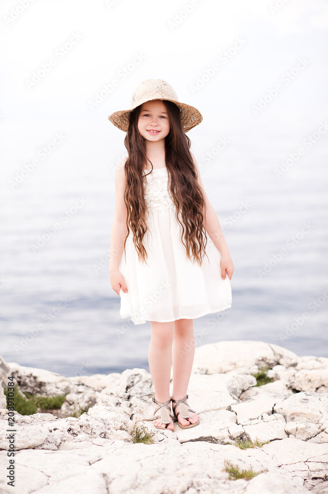 Stylish baby girl 4-5 year old wearing white dress and hat outdoors. Looking at camera. Summer time.