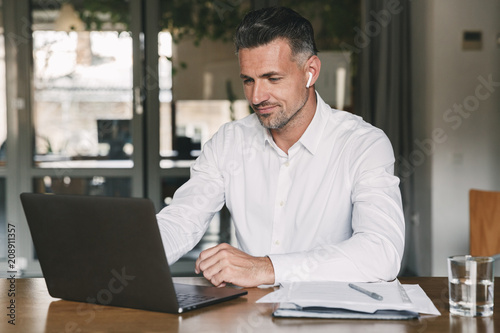 Image of handsome business man 30s wearing white shirt sitting at table in office, and working at laptop using wireless earpod