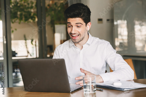 Image of cheerful office man wearing white shirt and modern earpod smiling while sitting at table in office, and speaking to laptop during video call or chat
