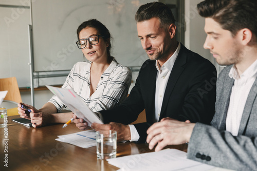 Group of business people in formal suits sitting at table in office, and examining resume of new personnel during job interview - business, career and placement concept
