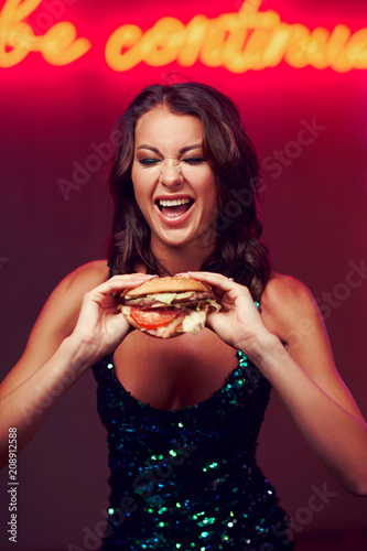 Sexy gorgeous woman in night dress with sequins eating hamburger in night club. Girl standing and posing against red wall with neon letters