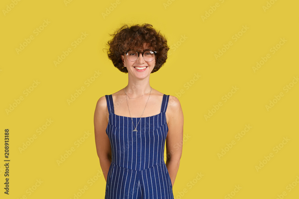 People, beauty and lifestyle concept. Attractive sensual girl with curly brown hair and wide smile dressed in blue overall smiling broadly on yellow background. Joyful nice female People, beauty and lifestyle concept. Attractive sensual girl with curly brown hair and wide smile dressed in blue overall smiling broadly on yellow background. Joyful nice female