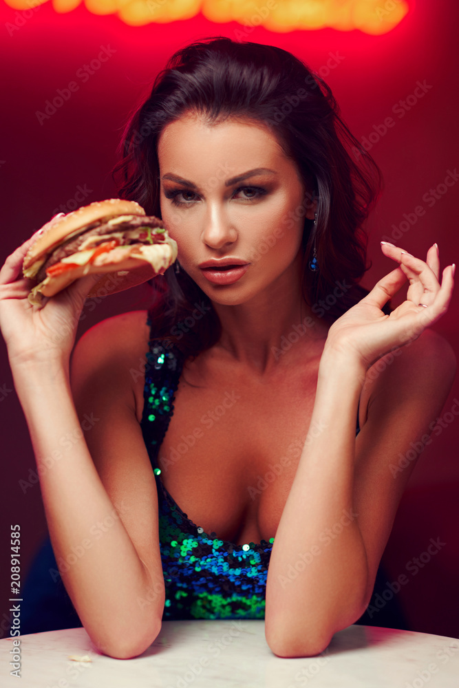 Sexy woman sitting at table in night club or bar and eating hamburger. Fashion style portrait Sexy woman sitting at table in night club or bar and eating hamburger. Fashion style portrait