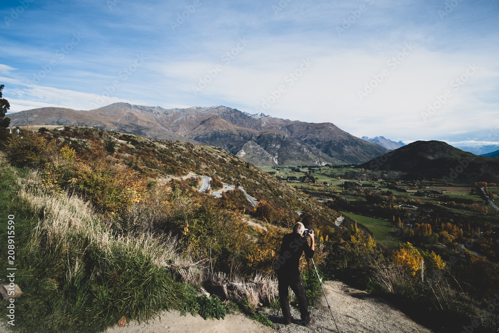 A man taking a photo of a beautiful road between Queenstown and Wanaka via Crown range. Grassland autumn trees with beautiful landscape of rocky mountains. dramatic effect. A man taking a photo of a beautiful road between Queenstown and Wanaka via Crown range. Grassland autumn trees with beautiful landscape of rocky mountains. dramatic effect.