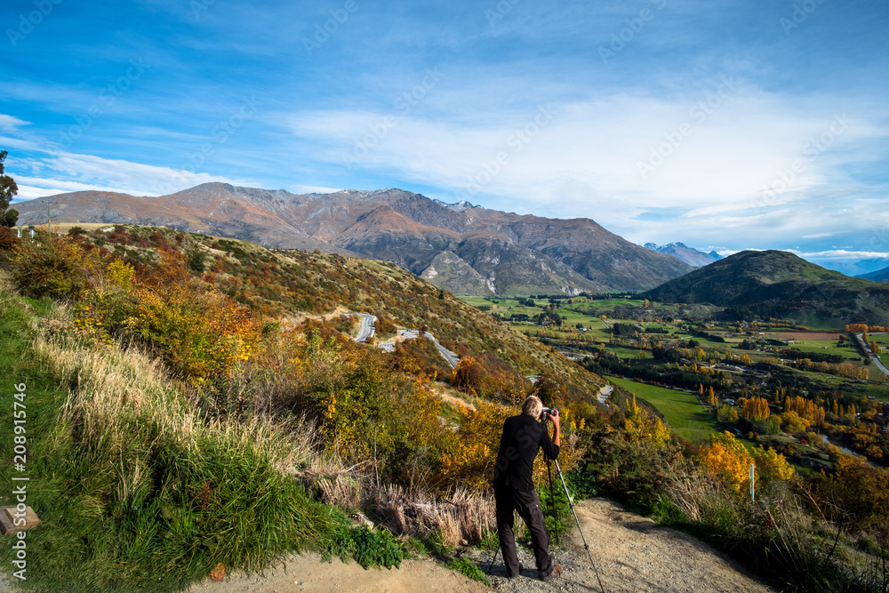 A man taking a photo of a beautiful road between Queenstown and Wanaka via Crown range. Grassland autumn trees with beautiful landscape of rocky mountains.