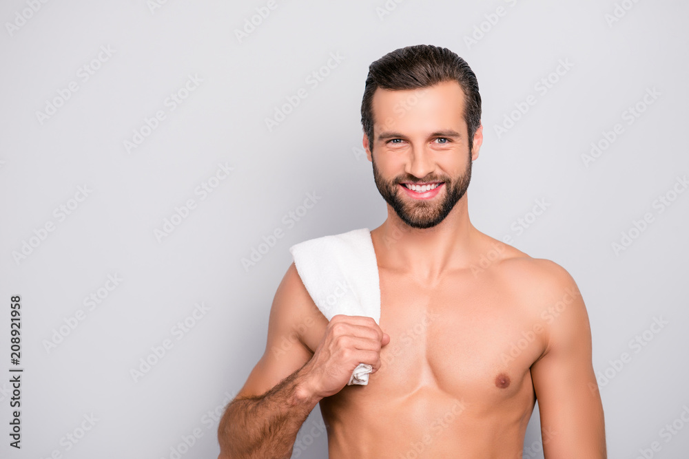 Fototapeta premium Portrait with copy space of brunet, cheerful man with beaming smile and bristle having a towel on his shoulder, holding towel with hand, looking happy after bath, standing over grey background
