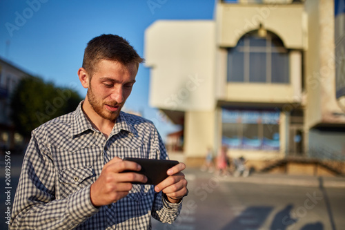 Half length portrait of bearded male in casual clothes using cell telephone while standing in urban setting.