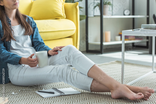 Canvas Print cropped shot of barefoot girl holding cup while sitting on carpet and studying a