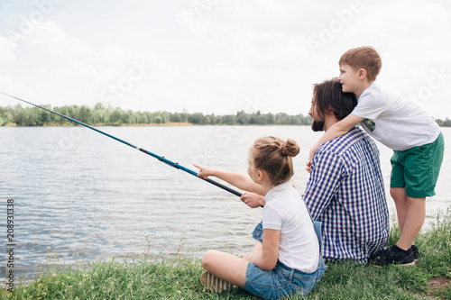 Dad and his children are spending time together at river shore. They are trying to catch some fish. Man is holding fish-rod in hands. Boy is standing behind him and hugging. Girl is pointing forward.