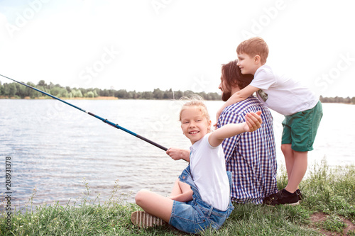 Man is sitting on grass and holding fish-rod. He is trying to catch some fish. Boy is standing behind and hugging him. Girl is looking behind and waving on camera.