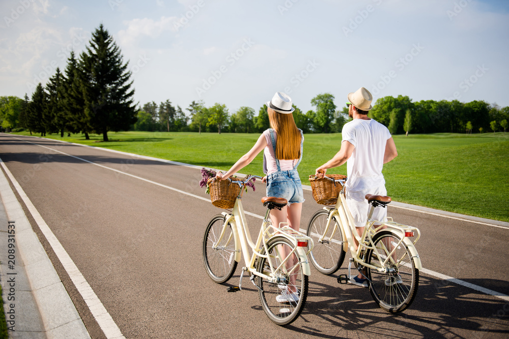 Rear view full-length portrait of active sportive couple in casual outfit straw hats shorts walking with retro bikes after riding over field forest. Sunny day daydream sunshine concept