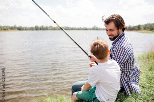 Bearded and handsome guy is sitting with his son at the edge of river shore and fishing. He is looking at son and smiling. He is proud of him.