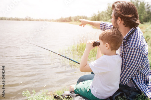Boy is sitting with his dad at the river shore and looking through binoculars. Adult man is pointing forward and holding fish-rod in right hand.