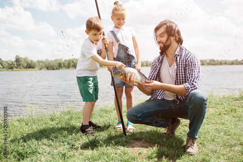 Funny picture of kids standing behind their father and looking at fish. They are amazed. Boy is holding fish-rod and touching fish. Guy is holding it and showing it to kids.