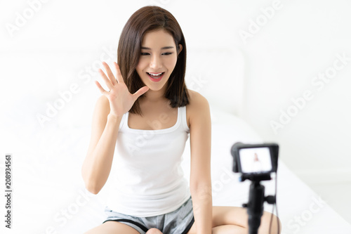 Young Asian woman blogger saying hi and waving with recording video sitting on white bedroom