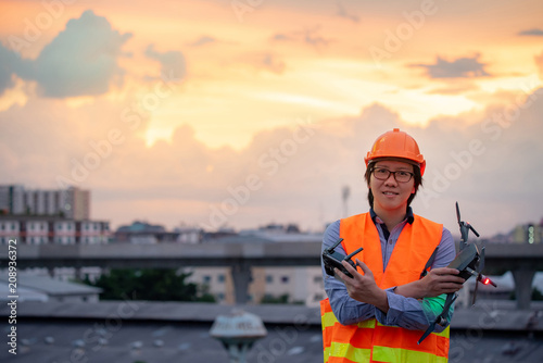 Young Asian engineer holding drone at construction site during sunset. Using unmanned aerial vehicle (UAV) for land and building site survey in civil engineering project.